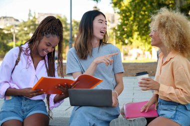 Üç mutlu çok ırklı arkadaş parkta dizüstü bilgisayar ve klasörle ders çalışıyor..