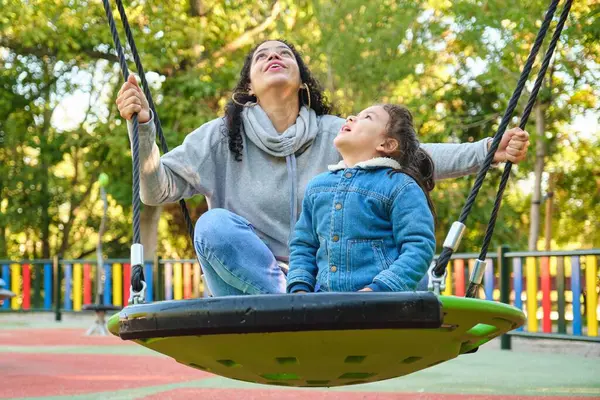 Happy Latin mother playing with her son looking up on a swing in a park. Latin family.