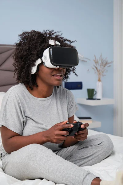 African young woman playing video games with a VR headset sitting on bed in the morning.