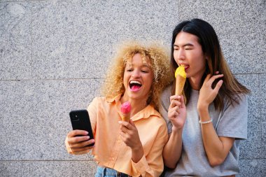 Dondurmalı iki çok ırklı mutlu arkadaş yazın caddede akıllı telefon kullanıyor..