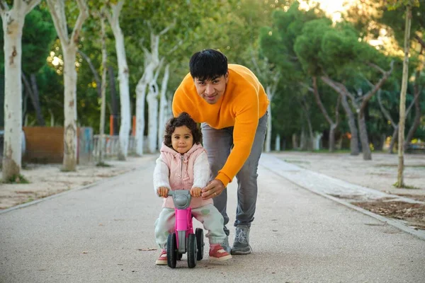 Latin father playing with his one year old daughter on a balance bike in a park.