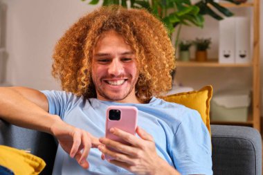 Genç mutlu Latin adam akıllı telefonunu evde kanepede yatarken kullanıyor..