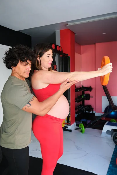 Pregnant woman strength exercising helped by the personal coach at gym.