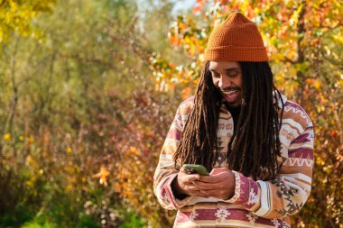 Güneşli bir sonbahar gününde akıllı telefonu kullanmaktan mutlu olan Afrikalı Amerikalı genç adam..