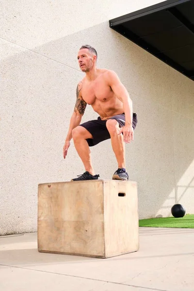 Strong man jumping on to wooden box from floor in cross training outdoor gym. CrossFit, fitness and sport.