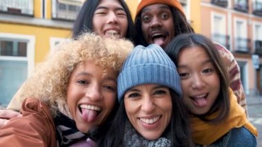 Çok ırklı arkadaş grubu, sonbaharda dışarıda akıllı telefonla selfie çekiyor. Mutlu gençler şehir caddesinde yürürken eğleniyor. Dışarıda eğlenen kız ve erkeklerle arkadaşlık kavramı..