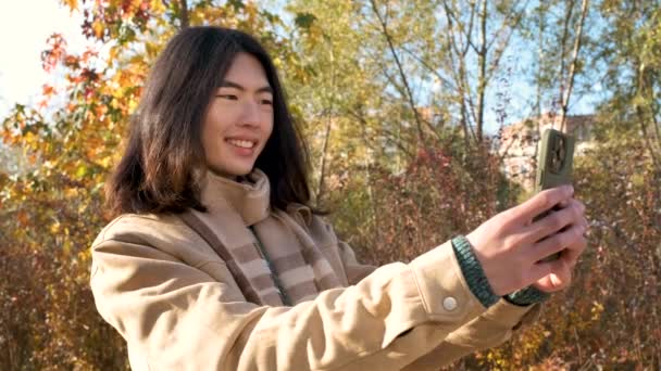 長い髪の若いアジアの男は 秋の公園で面白いセルフィーを取って幸せ — ストック動画