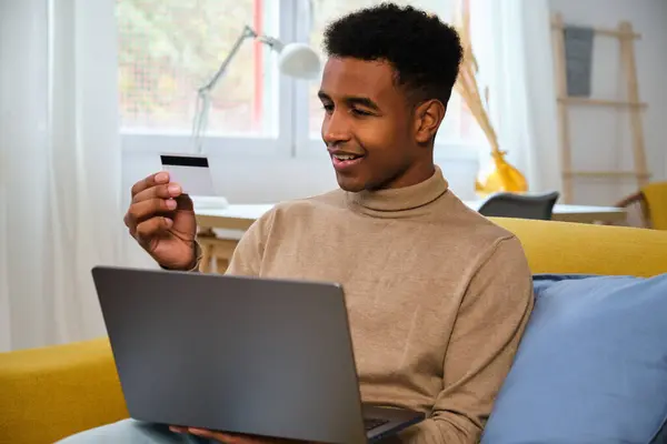 African young man buying online with a credit card in his laptop at home. Black Friday sales.