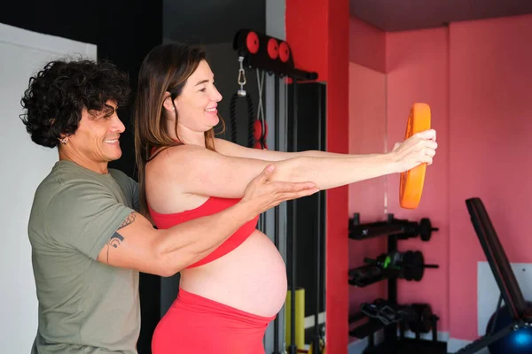 Pregnant woman strength exercising helped by the personal coach at gym.