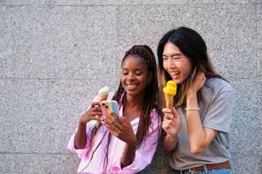 Dondurmalı iki çok ırklı mutlu arkadaş yazın caddede akıllı telefon kullanıyor..