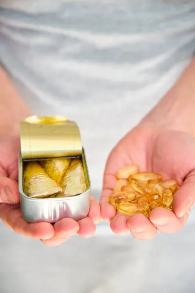 A person is holding a can of sardines in one hand and omega 3 fish oil capsules in the other.