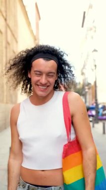 Beyaz kolsuz tişörtlü ve gökkuşağı çantalı eşcinsel adam Onur Yürüyüşü sırasında gülümsüyor ve kameraya bakıyor..