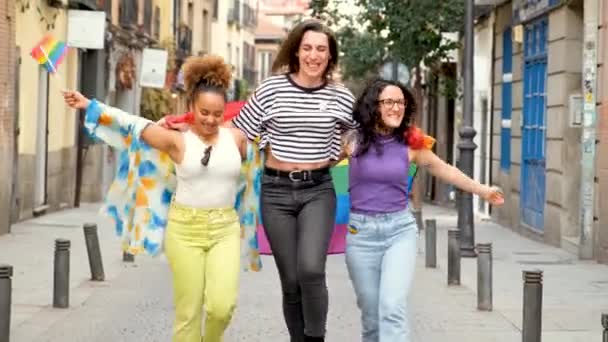 3人のレズビアンとトランスジェンダーの女性が 虹の旗に包まれた通りを歩いている シーンは楽しいし お祝いしています — ストック動画