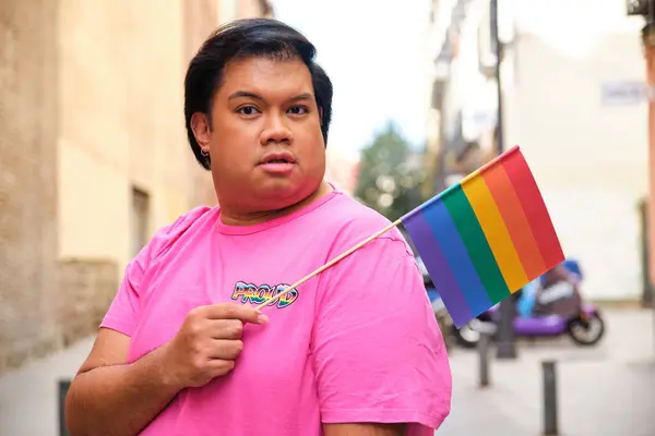 stock image Gay Asian man in a pink shirt holding a rainbow flag. He is looking at the camera. The flag is a symbol of the gay pride movement.