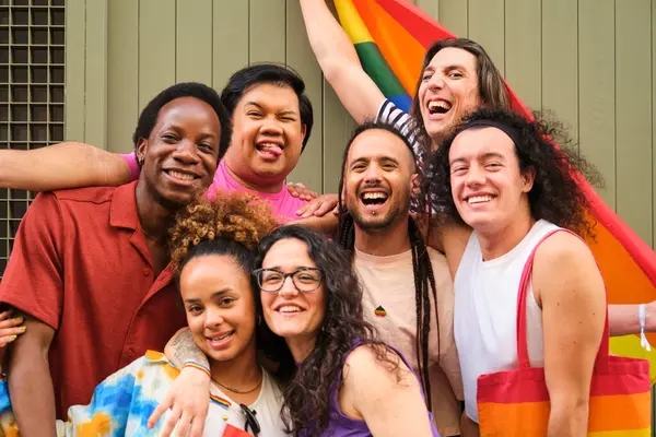 A group of people are posing for a picture with a rainbow flag at Pride Day. Scene is happy and celebratory.