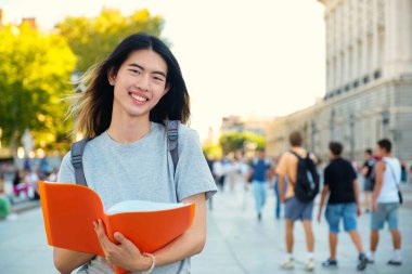 Tayvanlı erkek öğrenci İspanya 'da üniversite kampüsünde bir dosya ve sırt çantasıyla gülümsüyor ve kameraya bakıyor..