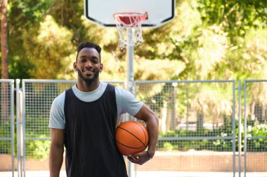 Atletik adam arka planda ağaçlar ve çitlerle basketbol sahasında poz veriyor.
