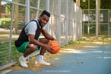 Atletik genç adam basketbol sahasında elinde basketbol topuyla çömeliyor.