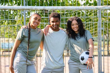 Üç çok ırklı futbolcu, bir futbol topuyla bir sahada yan yana duruyorlar.