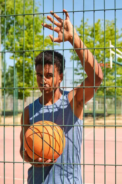 Güneşli bir günde çitin arkasında basket topu tutan genç bir adam.