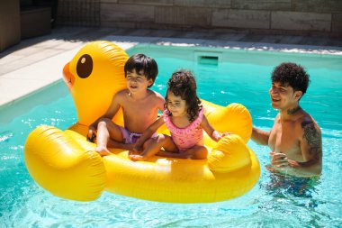 Bir adam çocuklarıyla havuzda sarı şişme bir ördeğin üstünde oynuyor. Sahne mutlu ve neşeli.