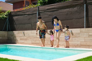 Dört kişilik bir aile el ele tutuşup havuza bakmaktan keyif alıyor. Kadın bikini giyerken, erkek şort giyiyor. Çocuklar da mayo giyiyor.
