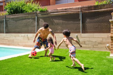 Bir adam ve iki çocuğu arka bahçede havuzun yanında futbol oynuyorlar. Adam ve çocuklar mayo giyiyor.