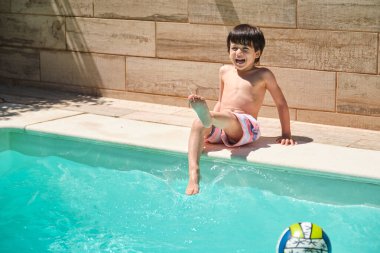 Genç bir çocuk havuzda oturmuş ayaklarıyla su sıçratıyordu. Gülümsüyor ve eğleniyor. Havuzda bir top var.