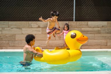 Babası ve kız kardeşiyle havuzda sarı şişme bir ördeğin üstüne atlayan bir çocuk. Baba şişme ördeği tutuyor ve kız kardeşi havuzun yanında duruyor. Sahne eğlenceli ve eğlenceli.