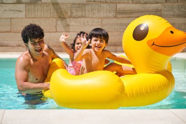 İki çocuk babalarıyla havuzda sarı şişme bir ördeğin üzerinde ellerini kameraya doğru sallıyor. Sahne neşeli ve neşeli.