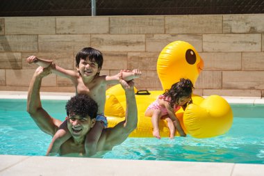Oğlu omuzlarında, kızı da havuzda sırtında bir adam. Kızı sarı şişme bir ördeğin üstünde.
