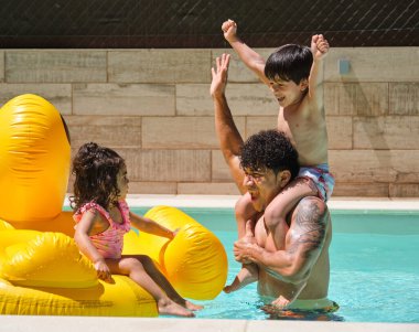 Oğlu omzunda bir adam ve kızı havuzda kutlama yapıyor. Çocuklar mayo giyiyor..