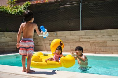Gülümseyen babasına ve kız kardeşine havuzda su püskürten bir çocuk. Kız kardeşi sarı şişme bir ördeğin üzerinde oturuyor.