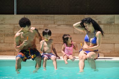 Dört kişilik bir aile havuzda oturmuş buzlu şeker yiyor ve birbirlerine bakıyorlar. Hepsi mayo giyiyor.