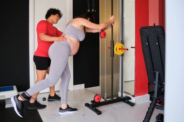 Spor salonunda bacak egzersizi yapan hamile bir kadına kişisel antrenör yardım ediyor..