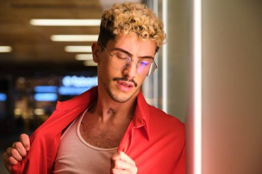 Bıyıklı, gözlüklü ve kırmızı gömlekli genç bir adamın portresi aydınlanmış bir duvara dayanıyor.