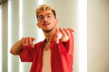 Saçları boyalı ve kırmızı elbiseli genç adam ışıklı bir koridorda dans ediyor.