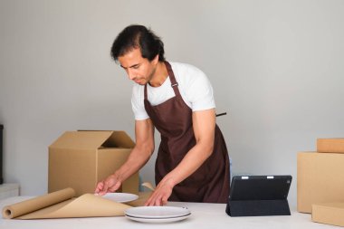 Karton kutuların yanında dijital tablet kullanan odaklanmış bir adam, teslimat için paket hazırlıyor