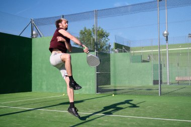 Genç adam güneşli bir yaz gününde kortta raket oynarken atlıyor.