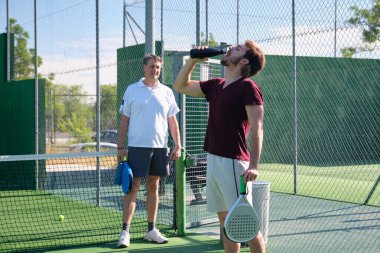 Koçla tenis oynadıktan sonra tekrar kullanılabilir şişeden su içen genç adam.