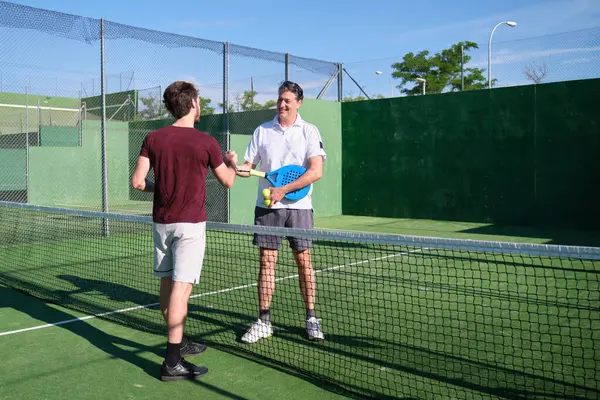 Güneşli bir günde oynanan maçtan sonra iki erkek raket tenisçi ağda el sıkışıyor.