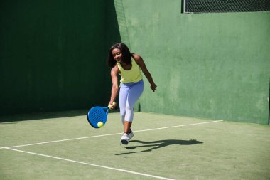Yeşil raket kortunda Padel oynayan genç bir kadın raketle topa vuruyor.