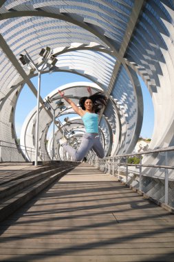 Genç bir kadın modern bir köprüde kolları havada havaya zıplıyor.