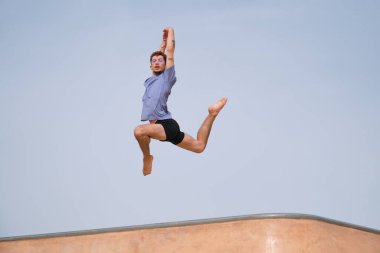 Mavi tişörtlü ve siyah şortlu genç adam zarif bir şekilde kaykay parkında havaya zıplıyor.