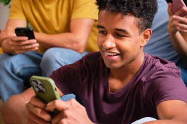 Genç siyahi bir adam akıllı telefonunu paylaşılan dairede arkadaşlarıyla otururken kullanıyor..