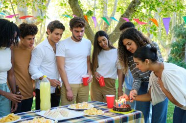 Bir grup arkadaş dışarıda bir piknik masasının etrafında toplanmış genç bir kadının doğum günü pastasının üzerindeki mumları yakmasını izliyorlar.