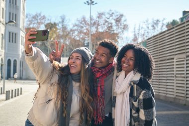 Sıcak kış kıyafetleri içinde üç genç etnik arkadaş dışarıda selfie çekerken parmaklarıyla zafer işareti yapıyorlar.