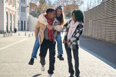 Üç neşeli çoklu etnik arkadaş sokakta yürüyor, biri diğerini sırtında taşıyor.