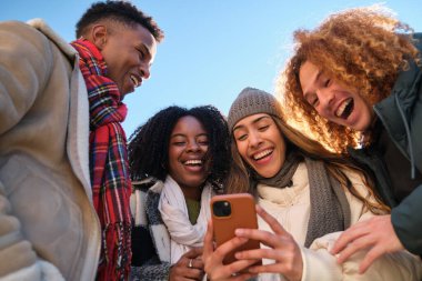 Dört neşeli genç öğrenci kış tatilinde akıllı telefon kullanıyor ve birlikte gülüyorlar.