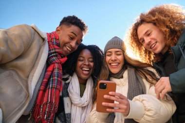 Dört neşeli çoklu etnik genç öğrenci kış boyunca akıllı telefondan komik bir şey izliyorlar.